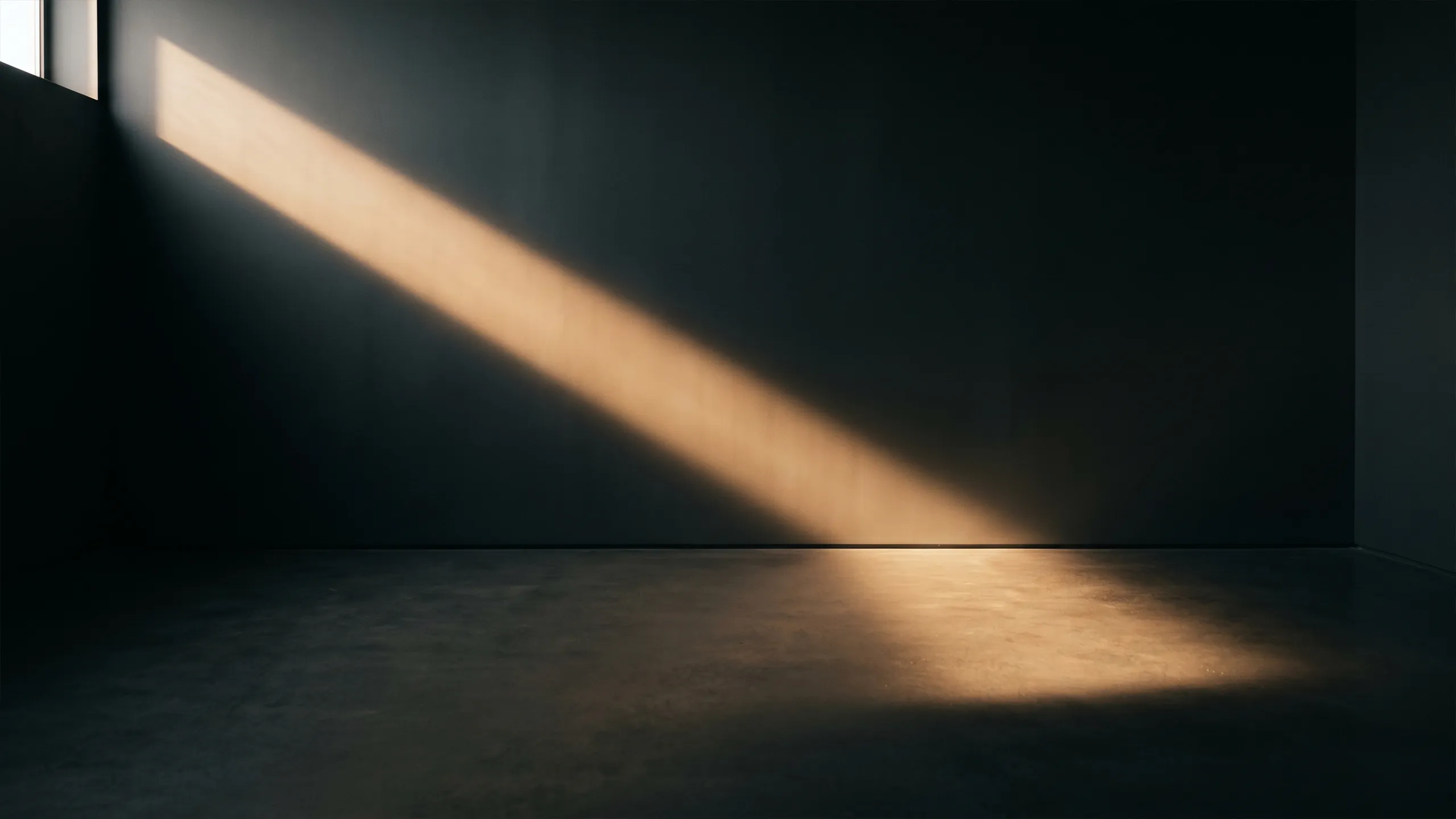 A diagonal beam of warm sunlight crossing a dark empty interior and revealing the texture of the wall and floor.