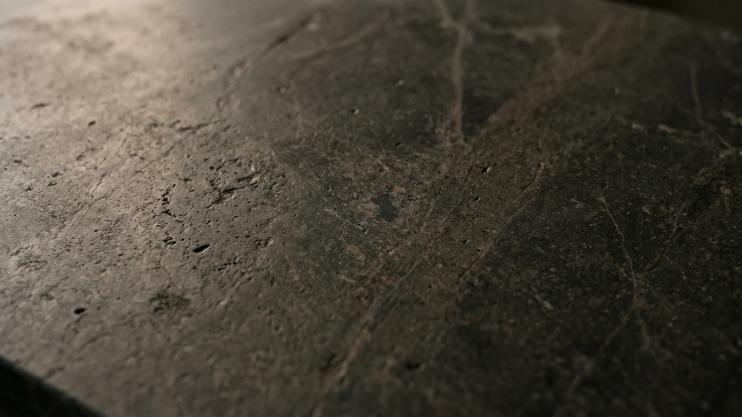 Close view of a dark weathered surface with pits, scratches and soft raking light revealing its material texture.